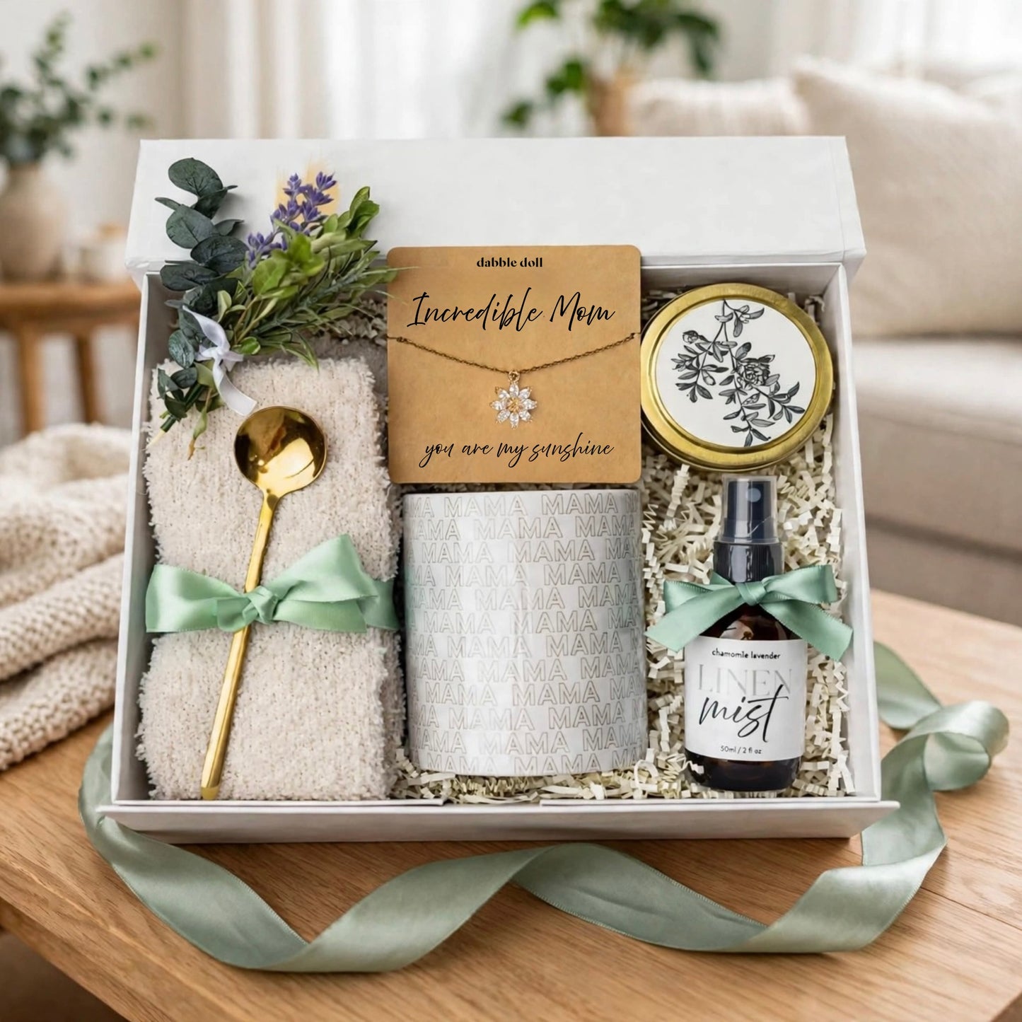 A curated gift box set on a wooden table, featuring a folded cream towel with a mint green ribbon, a gold spoon, a textured silver candle jar with a card reading 'Incredible Mom', a small amber spray bottle labeled 'Mist', and a round tin with botanical artwork. The box is styled with greenery and flowers, with a soft-focus living room background.