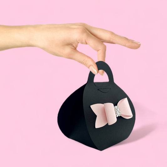 Hand holding black purse favor bags with pink bow and rhinestone detail against pink background