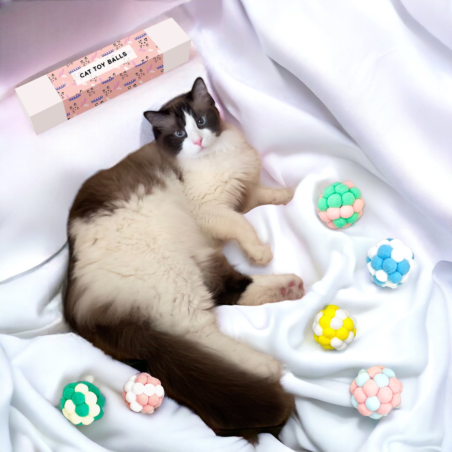 Cat lying on white fabric surrounded by colorful cat plush toy balls from Dabble Doll