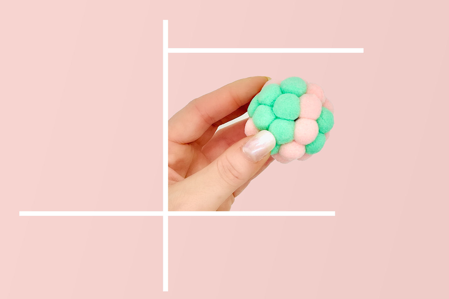 Hand holding a green and pink cat plush toy balls against a soft pink background