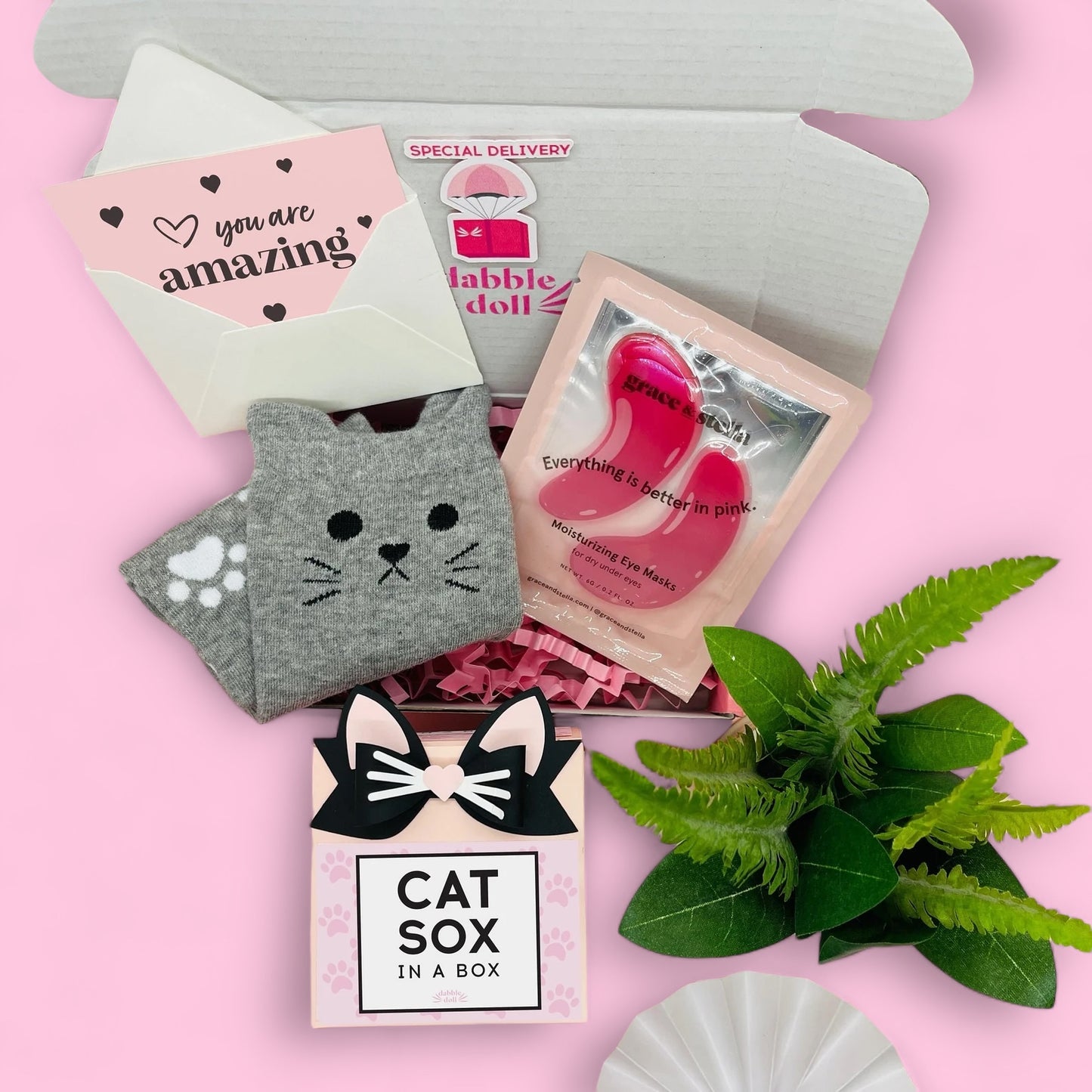 Dabble Doll cat socks gift box featuring gray cat-themed socks, pink moisturizing eye mask, and greeting card