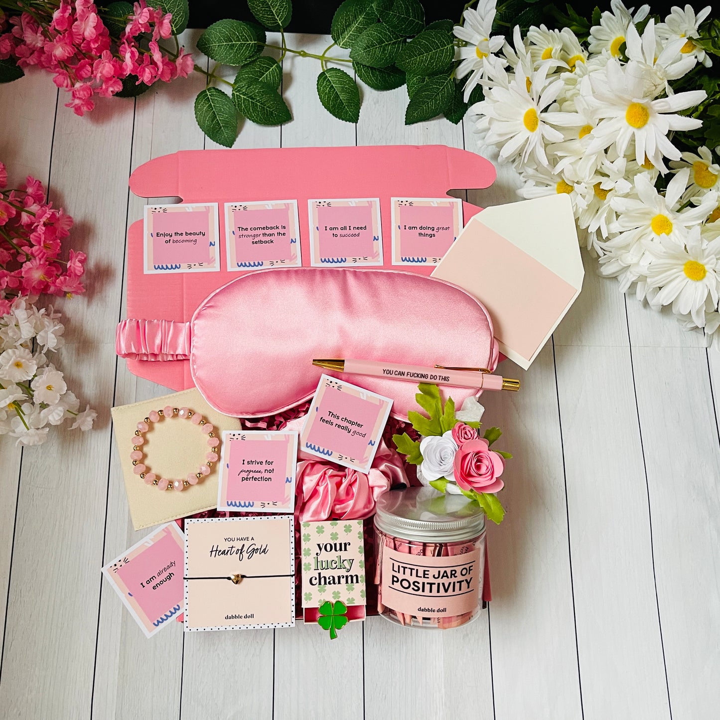Dabble Doll positivity care package gift box with pink eye mask, motivational cards, bracelet, flower, and positivity jar