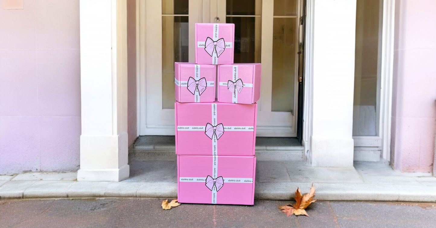 Stack of pink Dabble Doll thinking of you gift box for her with white ribbon bows in front of a doorway