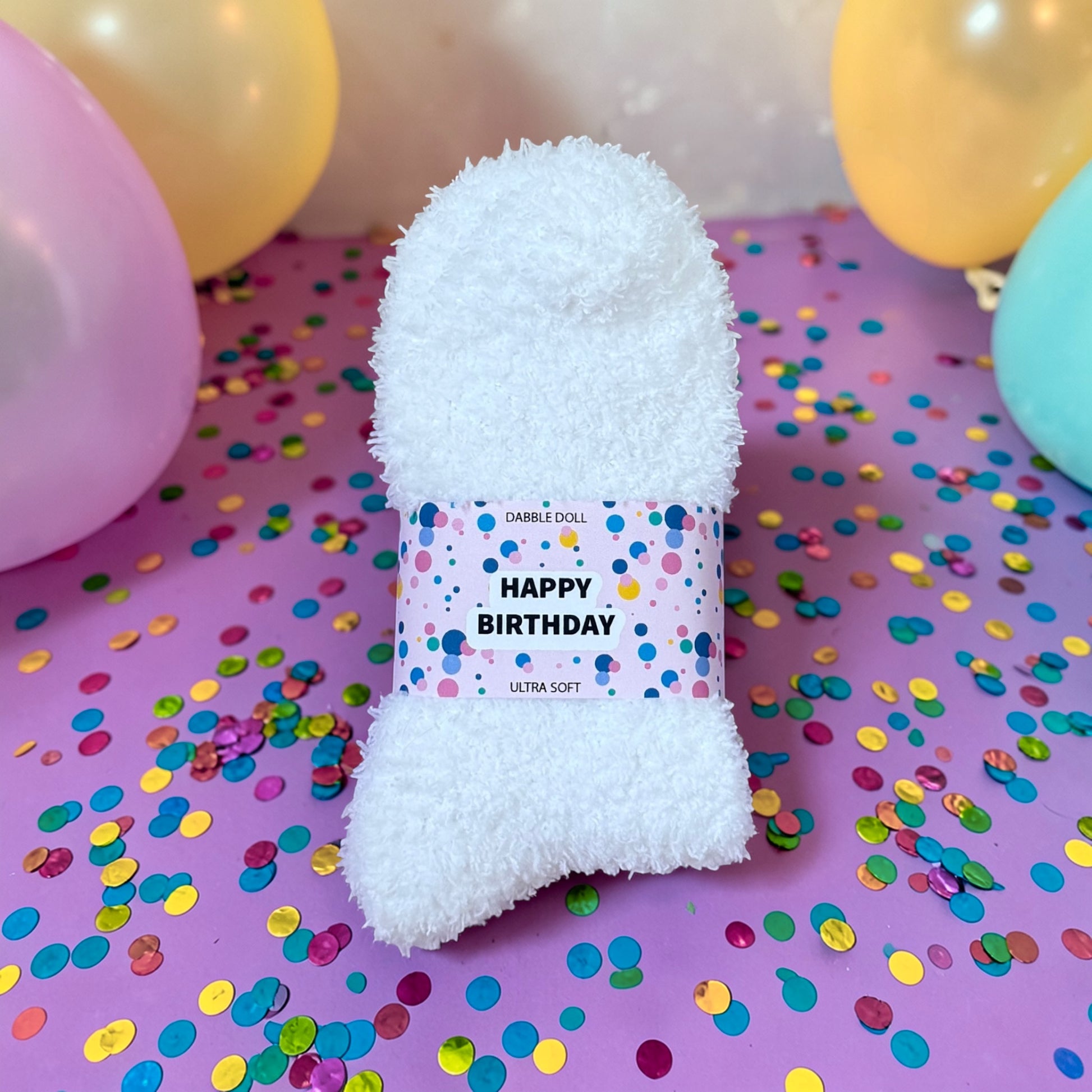 White fluffy toy with a 'Happy Birthday' label on a purple surface with colorful confetti and balloons.