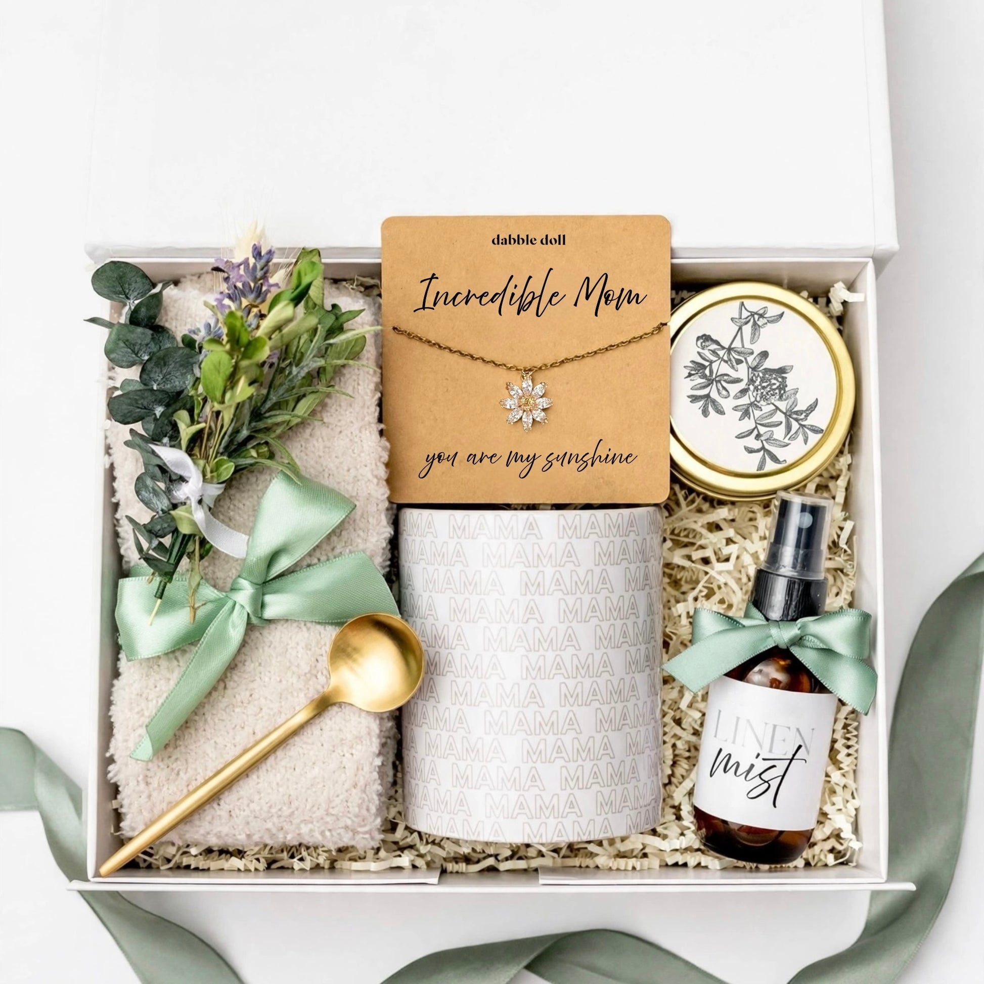 A curated gift box set for 'Incredible Mom' featuring a gold necklace with a flower pendant on a card, a white patterned mug, a beige towel with a sage green ribbon, a gold spoon, a small amber spray bottle, and a gold tin, all arranged in shredded paper inside a white box.