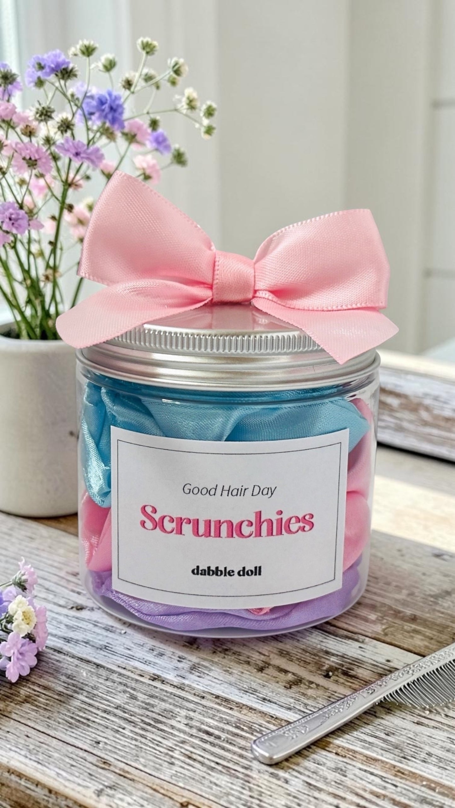 Jar of scrunchies with a pink bow on a wooden surface with flowers in the background