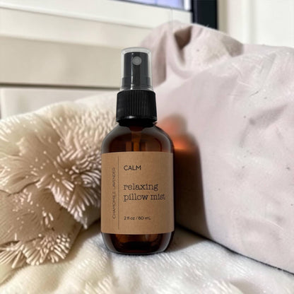 Brown spray bottle labeled 'Calm Relaxing Pillow Mist' on a textured surface with pillows in the background.