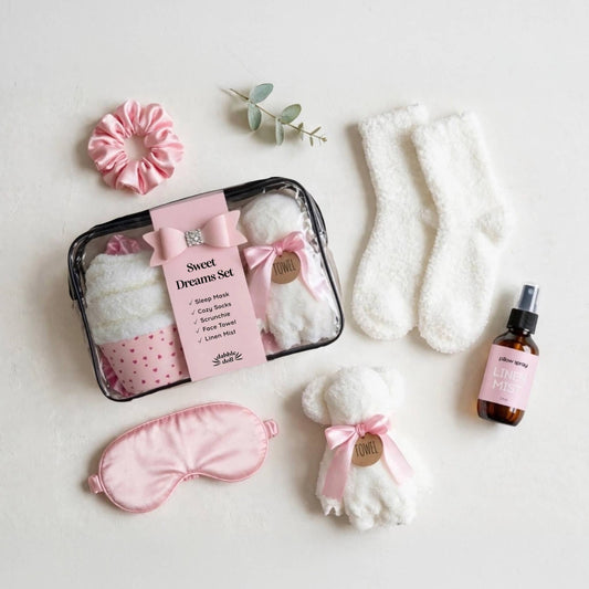 A flat-lay presentation of a 'Sweet Dreams Set' travel gift box containing a pink polka-dot sock, a white plush sock, a pink satin sleep mask, a white plush towel, a pink scrunchie, and a travel spray bottle, all arranged on a light cream background.