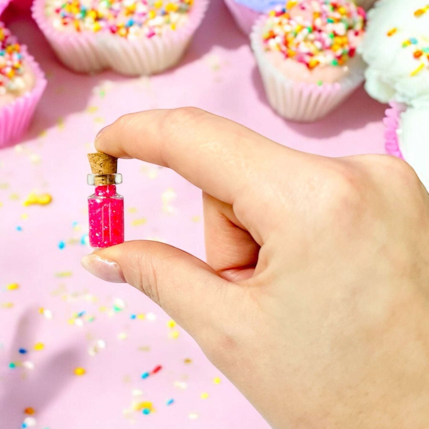 Dabble Doll Birthday Party Confetti in a Small Bottle Held by Hand with Colorful Cupcakes in Background