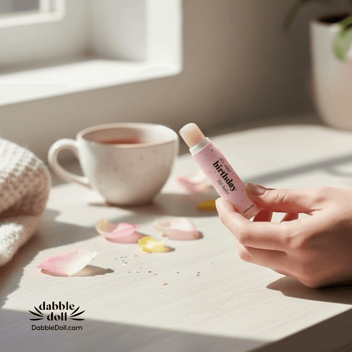 Dabble Doll Birthday Scented Lip Balm Held by a Hand Next to a Mug and Flower Petals