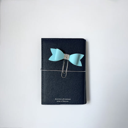 Dabble Doll Bow paperclip in light blue attached to a black notebook for stylish organization
