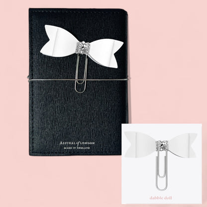 Dabble Doll Bow paperclip in white with rhinestone detail, accessorizing a black notebook
