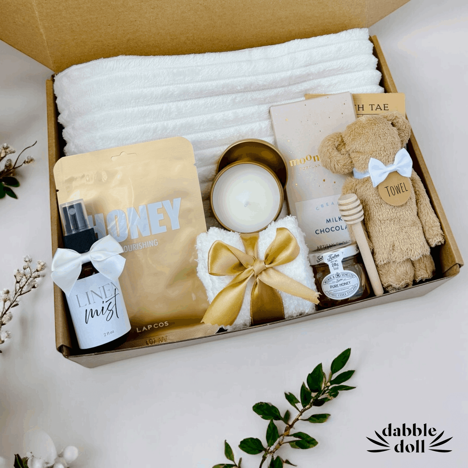 Dabble Doll gift in box featuring cozy towel, honey mask, candle, chocolate, and toy bear