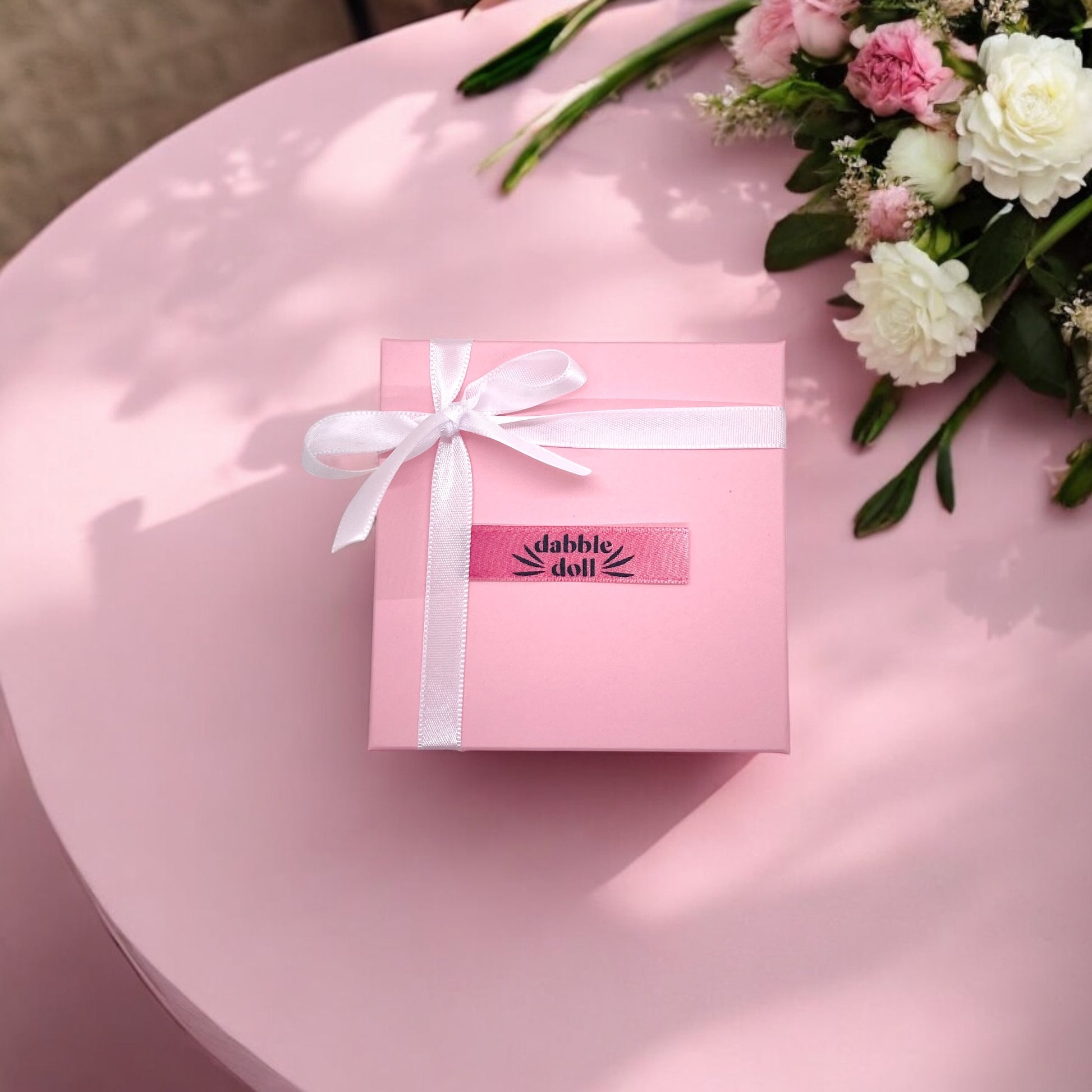 Pink gift box with white ribbon and Dabble Doll label on pink table near flowers