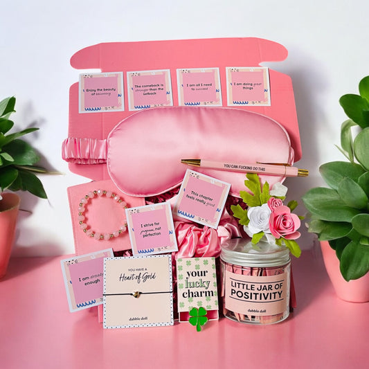 Dabble Doll positivity care package gift box with pink sleep mask, inspirational cards, bracelet, candle, and floral decor