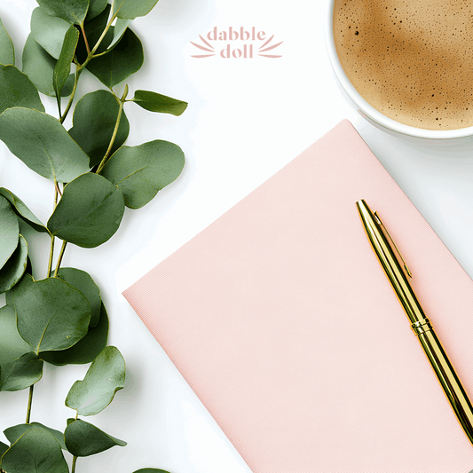Thoughtful hygge gift basket with Dabble Doll pink notebook, gold pen, green leaves, and cup of coffee