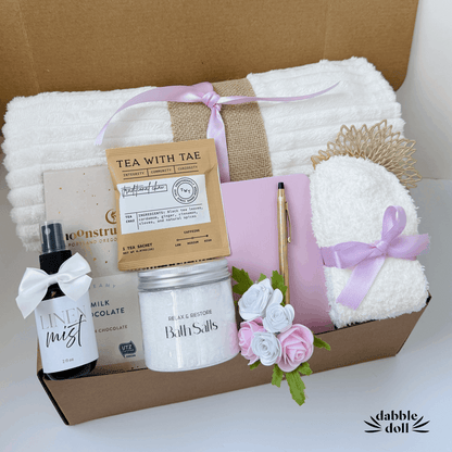 Thoughtful hygge gift basket by Dabble Doll with cozy towel, bath salts, tea sachet, linen mist, chocolate, and sleep mask